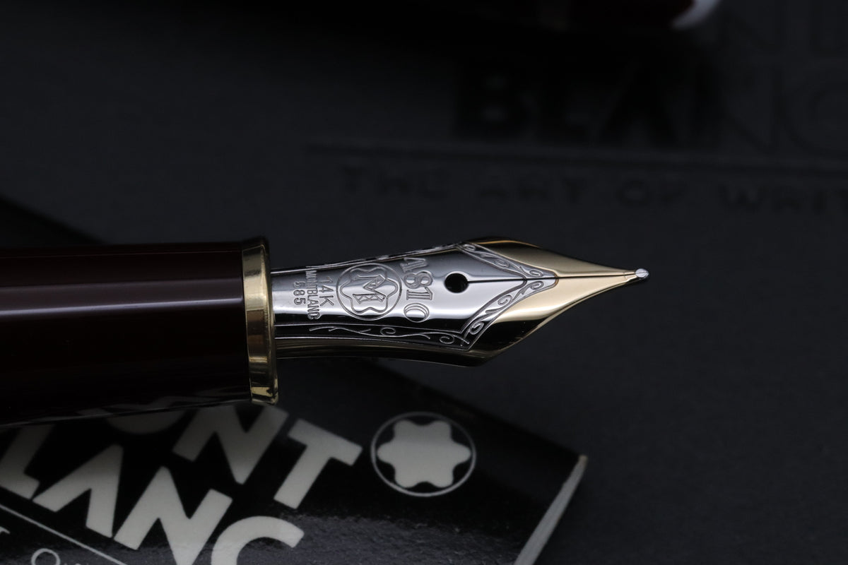 Close-up of a Montblanc fountain pen nib and upper barrel section, showing a polished silver nib engraved with the Montblanc mark and '14K' markings, with a dark reddish-brown barrel and gold trim, set against a dark background with a branded box partially visible.