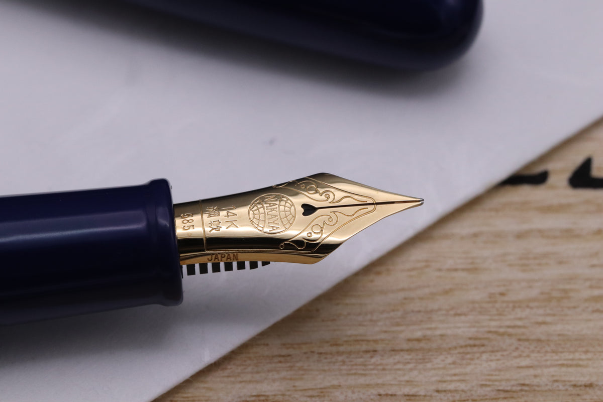 Close-up of a dark blue fountain pen nib and part of the cap resting on a white sheet of paper over a light wood surface.